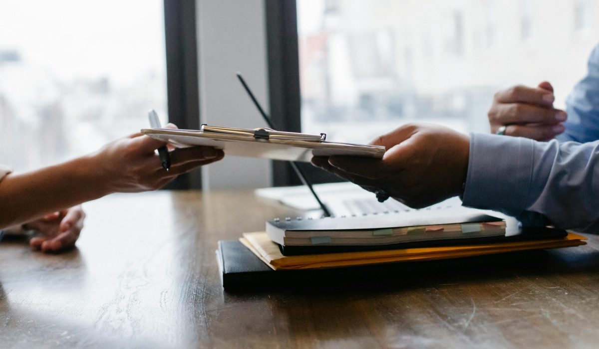 Crop anonymous ethnic woman passing clipboard to office worker with laptop during job interview